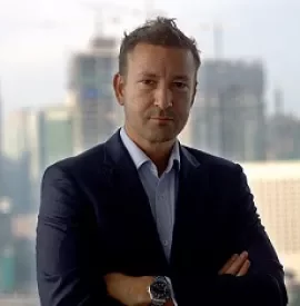 Man in suit standing with arms crossed, cityscape in background.