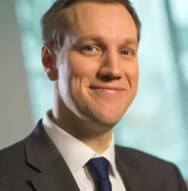 Smiling man in a suit with a blue tie, standing indoors.