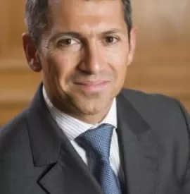 Smiling man in a suit with a blue tie, seated in a warmly lit room.
