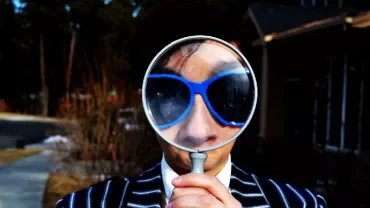 Person with sunglasses holding a magnifying glass in front of their face outdoors.