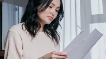 A woman reading a document next to a window with sheer curtains.