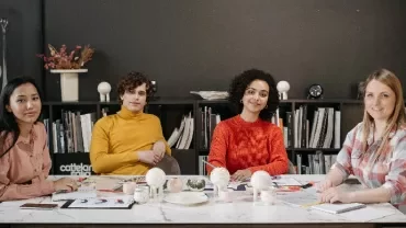 Four people sitting at a desk in an office with shelves in the background.
