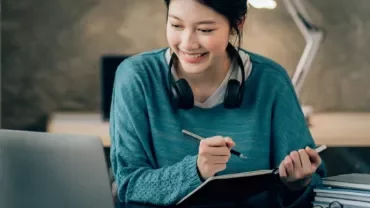 A woman with headphones writing in a notebook while looking at a laptop.