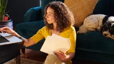 Woman works on laptop at home with dog sleeping on couch.