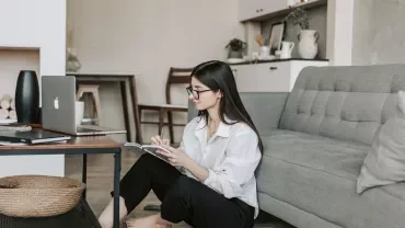 Woman sitting on the floor reading a magazine next to a sofa and laptop on a desk.