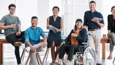 A diverse group of seven coworkers smiling in an office setting, gathered around a wooden table.