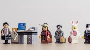 LEGO minifigures lined up, one in an office, others in costumes with briefcases.