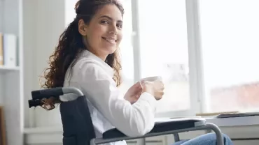 Woman in wheelchair smiling while holding a cup next to a laptop.