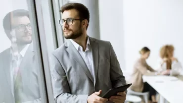 Man in suit holding a tablet, looking out the window, with colleagues working in the background.