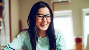 Woman with glasses smiling while using a laptop at home.