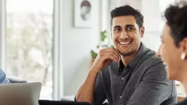 Group of people in a meeting, smiling and using laptops in a bright office space.