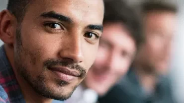 Man in a plaid shirt looking at the camera, with two others blurred in the background.
