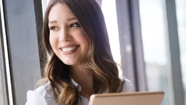 Smiling woman holding a tablet near a window with natural light.