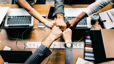 Five hands fist bump over a wooden table with laptops and coffee cups.