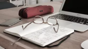 Open notebook with glasses on a wooden desk beside a laptop and red pencil case.