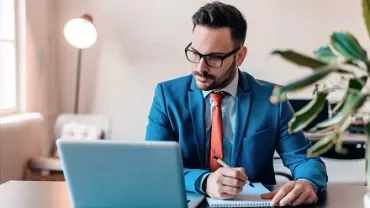 Man in a blue suit working on a laptop while taking notes.