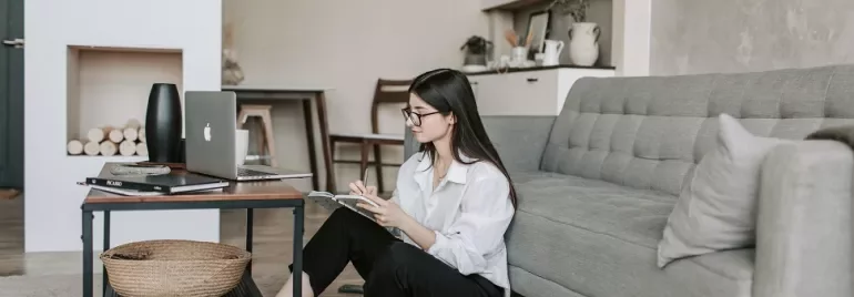 Woman sitting on the floor reading a magazine next to a sofa and laptop on a desk.