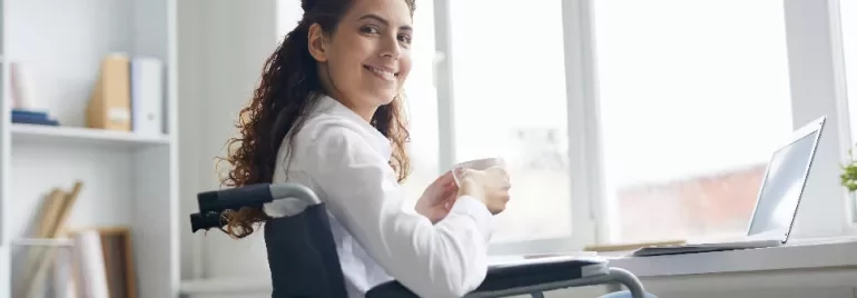 Woman in wheelchair smiling while holding a cup next to a laptop.