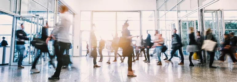 Blurred motion of people walking through a busy, bright lobby.