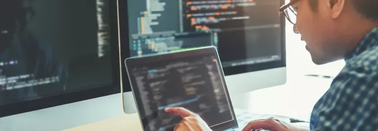 A person is working at a desk with a laptop and two large monitors displaying lines of code and programming software.