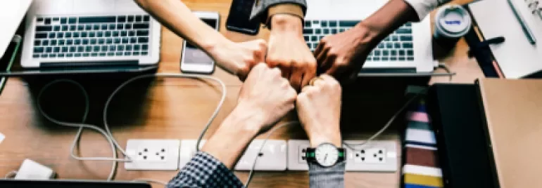 Five hands fist bump over a wooden table with laptops and coffee cups.