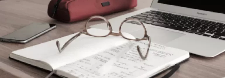 Open notebook with glasses on a wooden desk beside a laptop and red pencil case.