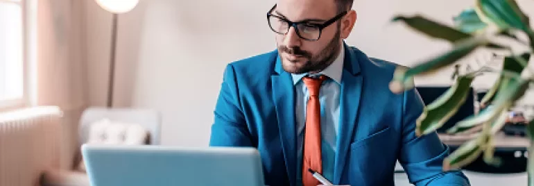 Man in a blue suit working on a laptop while taking notes.