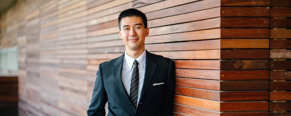 Smiling man in a suit leaning against a wooden wall.