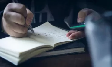 Person writing in a notebook while holding a phone, focused on taking notes.