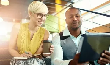 Two colleagues smiling while looking at a tablet. One holds a notebook and coffee cup.