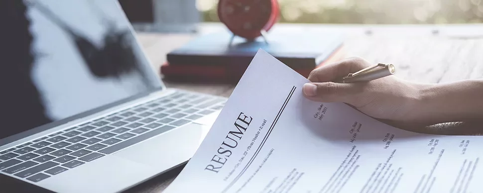 Person holding a resume near a laptop on a desk.