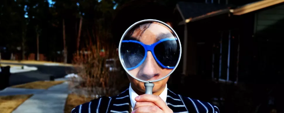 Person with sunglasses holding a magnifying glass in front of their face outdoors.