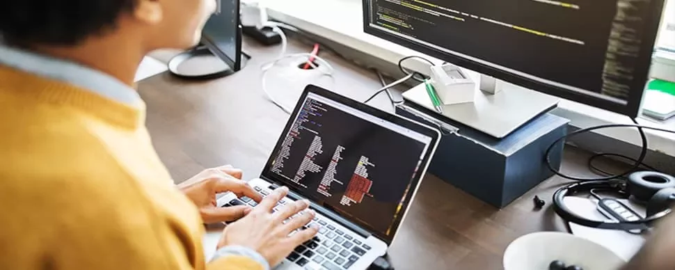 Person coding on a laptop with a large monitor displaying code.