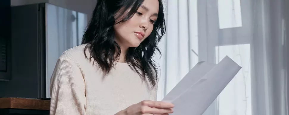 A woman reading a document next to a window with sheer curtains.