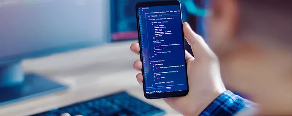 Person holding a smartphone displaying code on the screen, blurred computer monitor in the background.