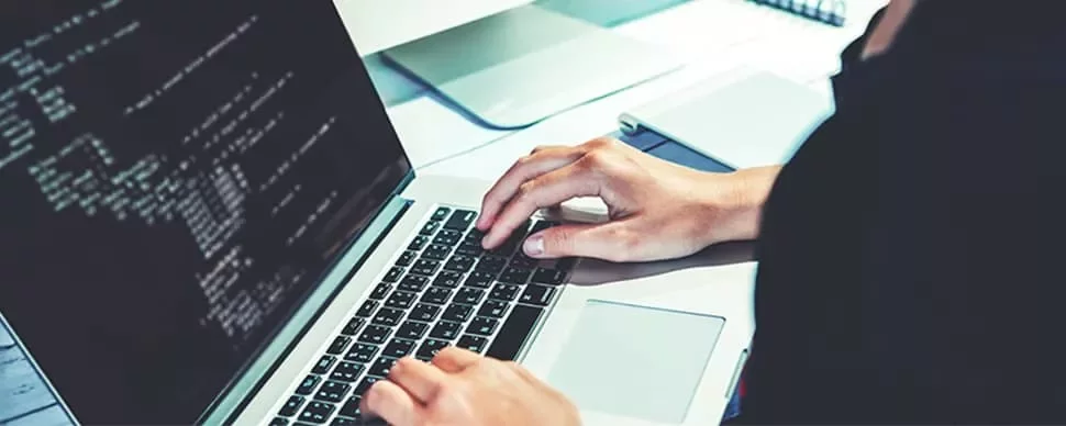 Person typing code on a laptop with a monitor displaying more code.