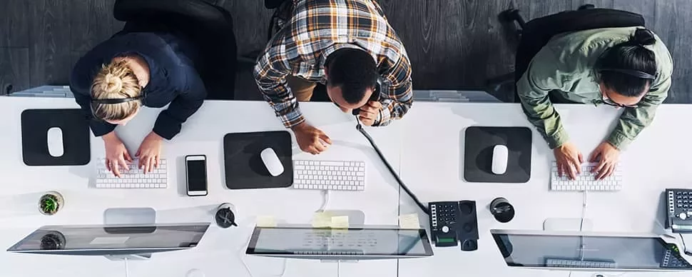 Overhead view of three people working at a desk with computers and headsets.