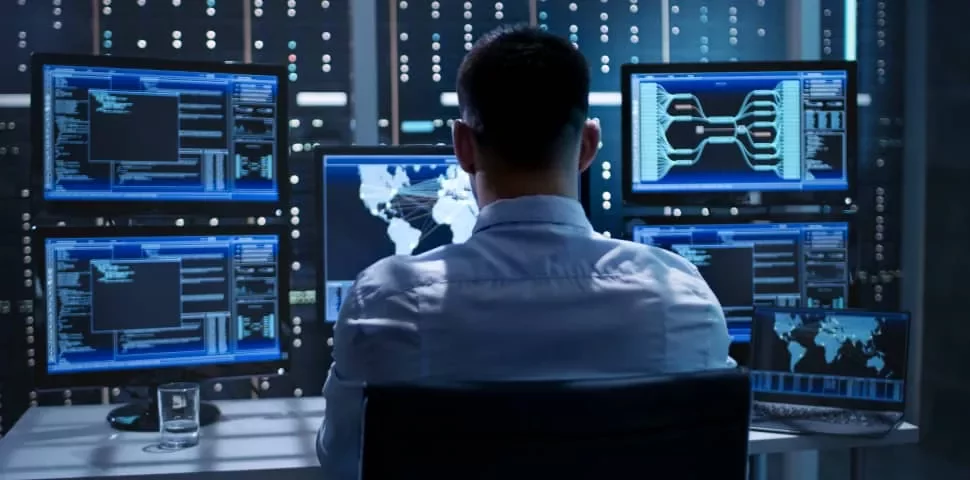 Person sitting at a desk, surrounded by multiple computer screens displaying data and maps.