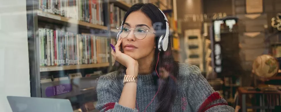 Woman in library wearing headphones, looking thoughtful.
