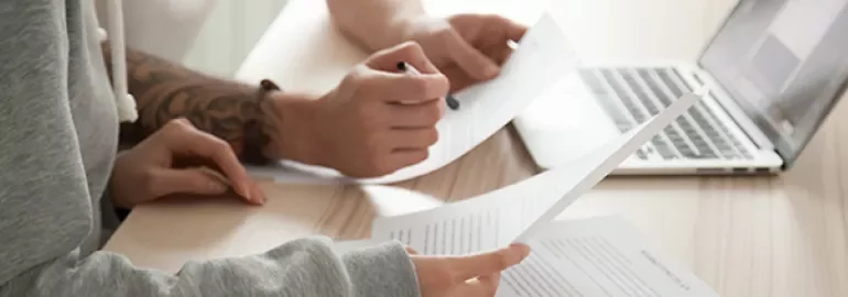 Two people reviewing documents with a laptop on a desk.