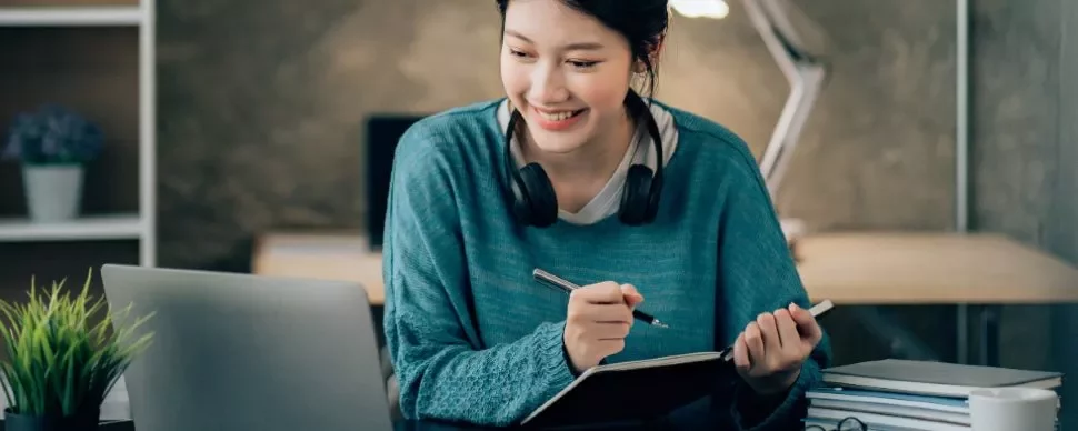 A woman with headphones writing in a notebook while looking at a laptop.