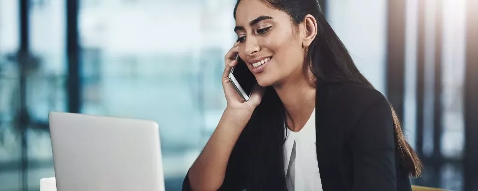 Smiling woman talking on phone while working on a laptop.