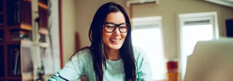 Woman with glasses smiling while using a laptop at home.