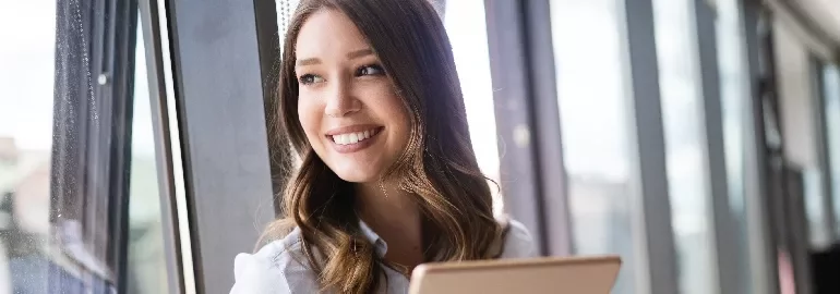 Smiling woman holding a tablet near a window with natural light.