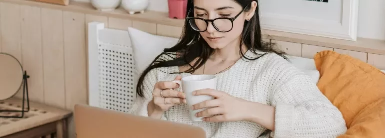 Woman with glasses sipping coffee, using a laptop on a cozy couch.