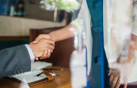 Two people shaking hands over a desk in an office setting.