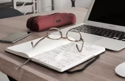 Open notebook with glasses on a wooden desk beside a laptop and red pencil case.