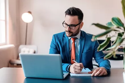 Man in a blue suit working on a laptop while taking notes.